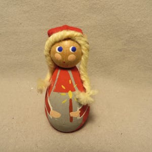 May include: A wooden doll with a round body and a painted face. The doll has blonde yarn hair, blue eyes, and a red hat. The doll's outfit is red and gray with a candle design. A decorative item.