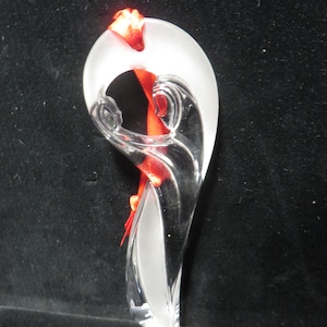 May include: A clear glass ornament with a frosted white top and a curved, flowing design. A red ribbon is threaded through the ornament. The ornament has a modern, abstract aesthetic.