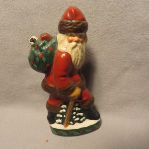 May include: A ceramic Santa Claus figurine, painted in red, brown, and white. Santa is carrying a green sack with red berries and a walking stick. The base is decorated with holly and snow.