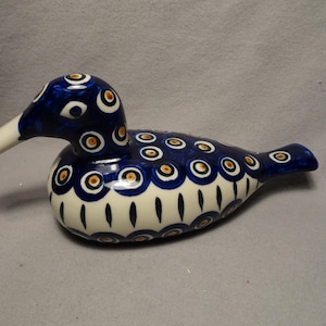 May include: A ceramic bird figurine featuring a glossy finish with a deep blue and cream color palette. The body is decorated with repeating circular peacock-eye patterns in blue, white, and orange, while the lower section displays vertical markings.