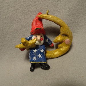 May include: A hand-painted ornament featuring a gnome wearing a red hat and a blue robe with white stars, holding a yellow star. The gnome is positioned next to a large, golden crescent moon with a smiling face and a small loop for hanging.