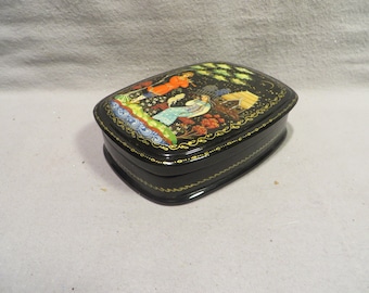 May include: Black lacquered wooden box with a painted scene of two figures in a forest setting. The box has a gold trim and a hinged lid.