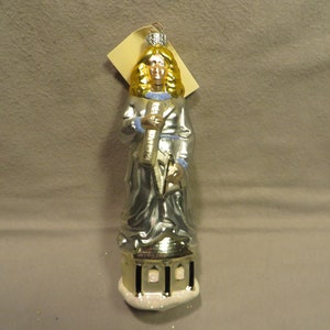 May include: Silver-colored glass ornament depicting a robed figure with golden hair, holding a scroll. The figure stands atop a building with black columns and a snowy base. A tag hangs from the top.