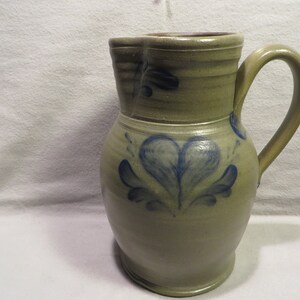 May include: A stoneware pitcher with a blue and white heart design. The pitcher is glazed in a light brown color and has a rounded body and a handle.
