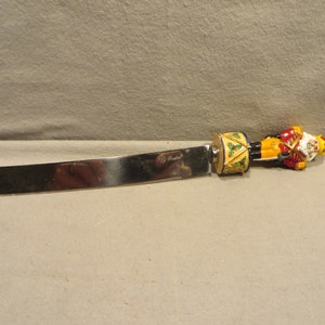 May include: A silver butter knife with a nutcracker handle. The handle is decorated with a yellow, red, and blue nutcracker figure playing a drum. The nutcracker is wearing a red and gold uniform with a blue sash. The handle is also decorated with green holly leaves.