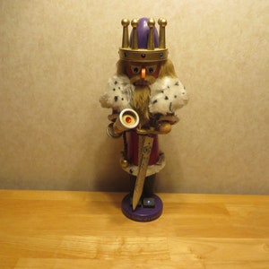 May include: A wooden nutcracker figurine of a king wearing a purple and gold crown, a white fur-trimmed robe, and holding a sword and a trumpet. The base of the nutcracker is purple and reads "A Soldier of Grace".