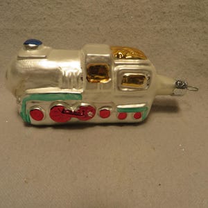 May include: A white glass Christmas ornament shaped like a train with red, green, and gold accents. The train has a blue chimney and a silver hook for hanging.