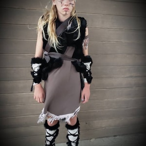 May include: A child in a costume, wearing a white lace headband, black and brown vest, and a gray skirt with a frayed hem. The costume includes black and white leg warmers with black ties and brown boots. The child has face paint.