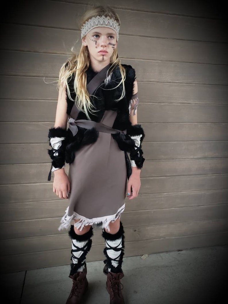 May include: A child in a costume, wearing a white lace headband, black and brown vest, and a gray skirt with a frayed hem. The costume includes black and white leg warmers with black ties and brown boots. The child has face paint.