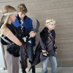 May include: Three children in fantasy costumes. The costumes include capes, faux fur trim, and decorative headbands. One child holds a curved, black prop weapon. The outfits are gray, brown, and blue.