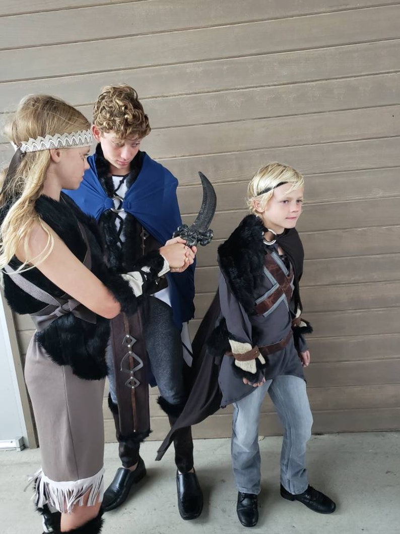 May include: Three children in fantasy costumes. The costumes include capes, faux fur trim, and decorative headbands. One child holds a curved, black prop weapon. The outfits are gray, brown, and blue.