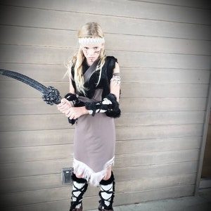 May include: A child in a warrior costume, holding a black sword. The outfit includes a gray dress with white fringe, black fur accents, and brown boots. A white headband and face paint complete the look. The background is a wooden wall.