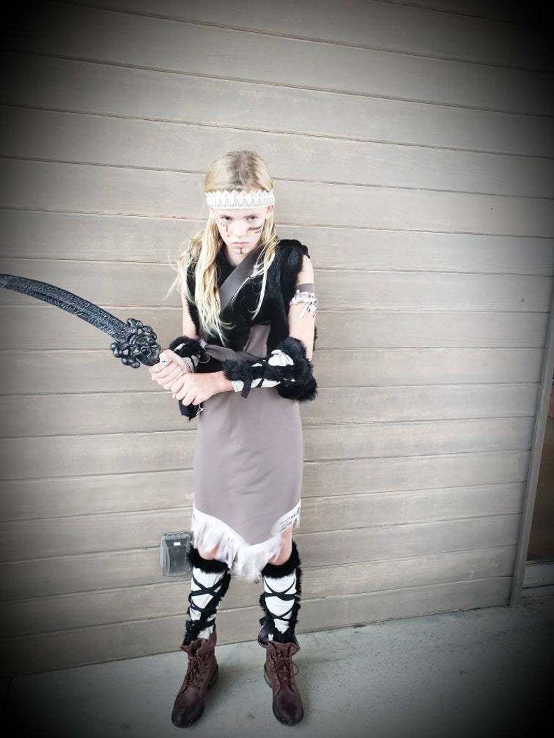 May include: A child in a warrior costume, holding a black sword. The outfit includes a gray dress with white fringe, black fur accents, and brown boots. A white headband and face paint complete the look. The background is a wooden wall.
