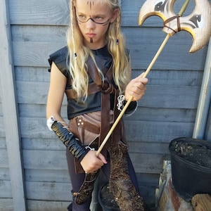 May include: A child in a warrior costume, holding a gold-colored axe. The costume includes a black top, brown leather accents, and matching leggings and boots. The child has face paint and long blonde hair.