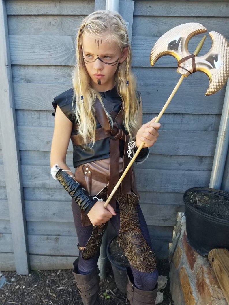 May include: A child in a warrior costume, holding a gold-colored axe. The costume includes a black top, brown leather accents, and matching leggings and boots. The child has face paint and long blonde hair.