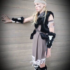 May include: A child's Native American-inspired costume featuring a brown dress with white fringe, a black fur vest, and matching arm and leg warmers. The outfit includes a beaded headband and face paint.