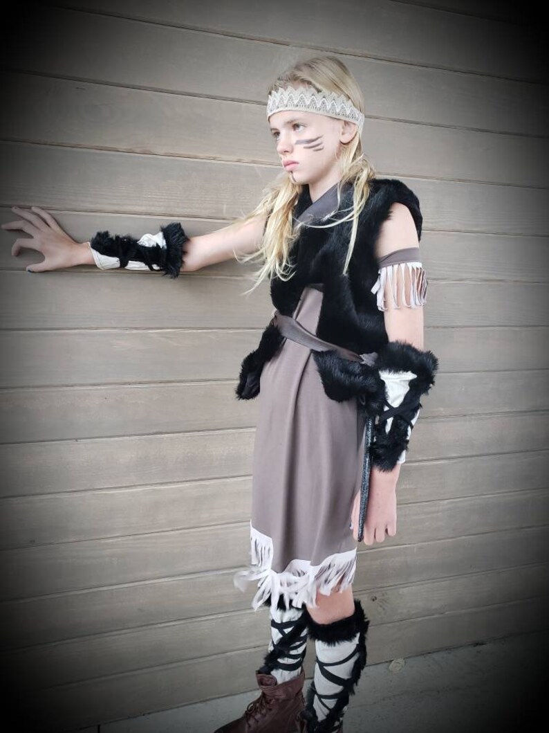 May include: A child's Native American-inspired costume featuring a brown dress with white fringe, a black fur vest, and matching arm and leg warmers. The outfit includes a beaded headband and face paint.
