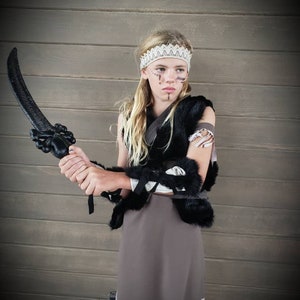 May include: A child in a costume, holding a black prop sword. The outfit includes a black fur vest, a gray skirt with white fringe, and a white lace headband. Face paint accents the look, creating a warrior theme.