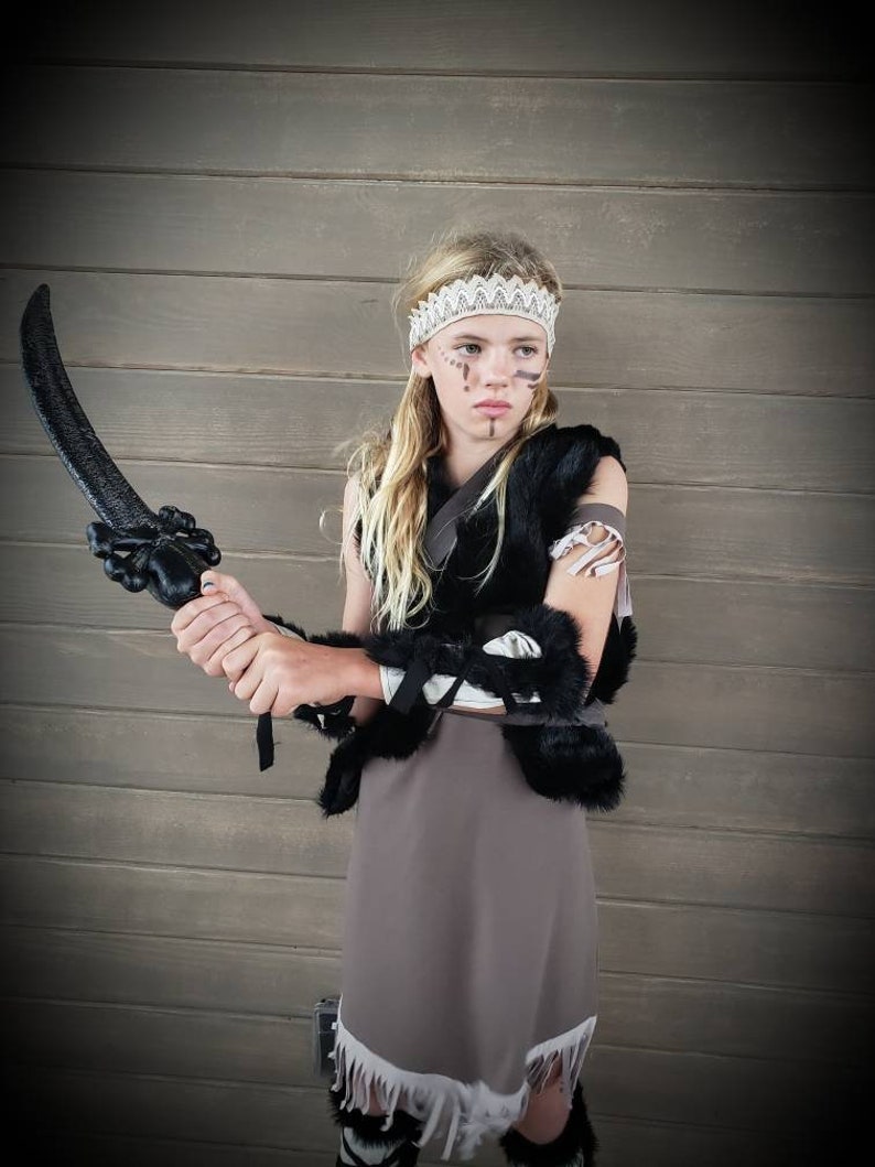 May include: A child in a costume, holding a black prop sword. The outfit includes a black fur vest, a gray skirt with white fringe, and a white lace headband. Face paint accents the look, creating a warrior theme.