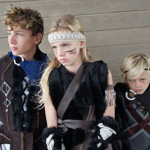 May include: Three children in costume, posing in front of a wooden wall. The costumes include faux fur, leather, and decorative metal rings. One child wears a blue scarf, another a headband, and a third a black cape. Face paint is visible on one child.