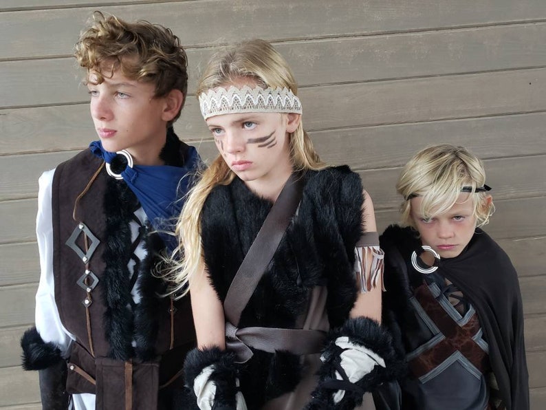 May include: Three children in costume, posing in front of a wooden wall. The costumes include faux fur, leather, and decorative metal rings. One child wears a blue scarf, another a headband, and a third a black cape. Face paint is visible on one child.