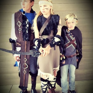 May include: Three children in fantasy costumes stand together. The costumes include faux fur, leather, and decorative elements. One child holds a sword. The outfits appear to be inspired by medieval or fantasy themes, with a focus on earthy tones and textures.