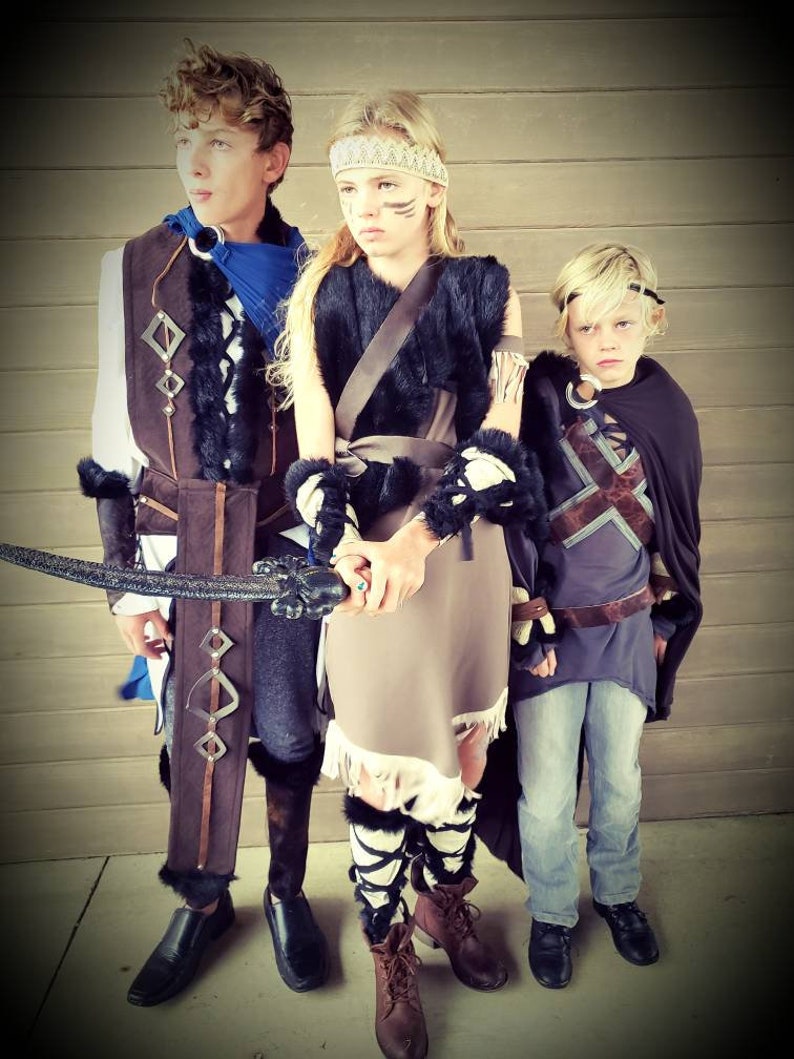 May include: Three children in fantasy costumes stand together. The costumes include faux fur, leather, and decorative elements. One child holds a sword. The outfits appear to be inspired by medieval or fantasy themes, with a focus on earthy tones and textures.