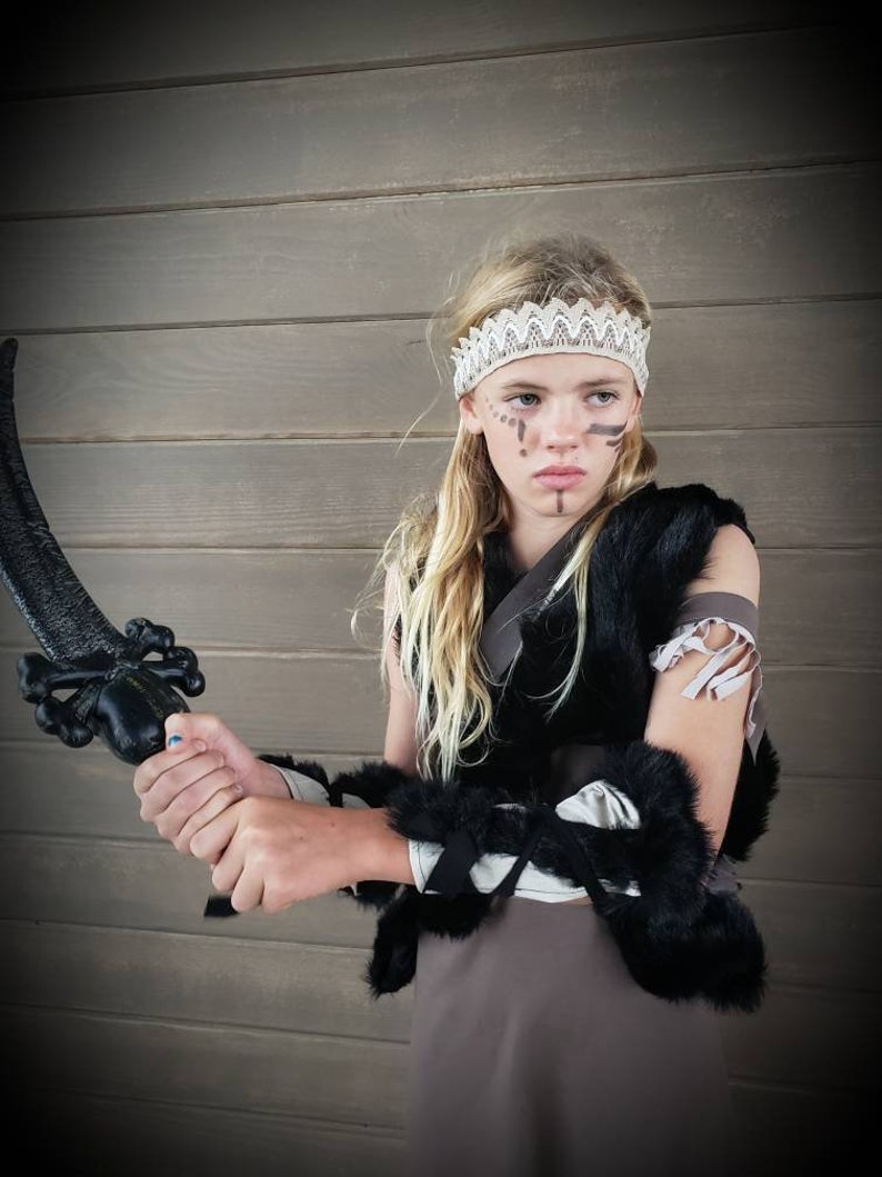 May include: A child in a warrior costume, holding a black sword. The outfit includes a fur vest, armbands, and a lace headband. The child has face paint and is standing in front of a wooden wall.