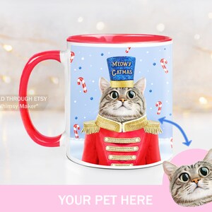 May include: A white ceramic mug with a red handle and rim. The mug features a cartoon cat dressed as a Christmas toy soldier with the text "MEOWY CATMAS". The image is a template for a personalized mug with a space for a photo of a pet.