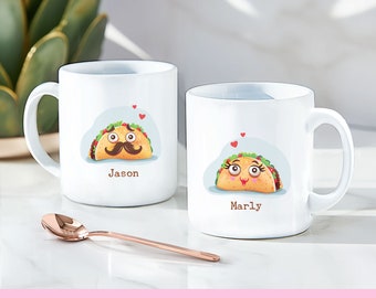 May include: Two white ceramic mugs with personalized names "Jason" and "Marly" featuring cartoon taco illustrations with facial expressions and hearts. The mugs are on a white surface with a rose gold spoon.