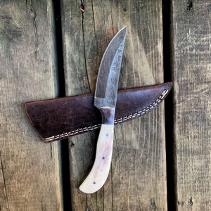 May include: A handcrafted hunting knife with a patterned steel blade and a bone handle. The knife rests on a brown leather sheath with white stitching, set against a wooden background. The blade is approximately 7 inches long.