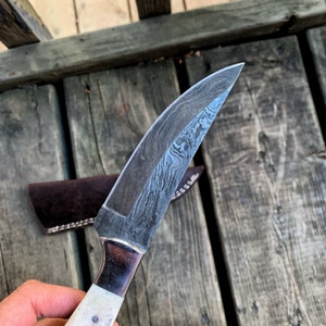 May include: A Damascus steel knife with a curved blade and a white handle. The blade has a unique pattern, and the handle is secured with a metal bolster. A brown leather sheath is visible in the background.