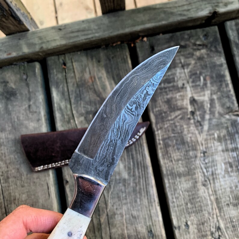 May include: A Damascus steel knife with a curved blade and a white handle. The blade has a unique pattern, and the handle is secured with a metal bolster. A brown leather sheath is visible in the background.