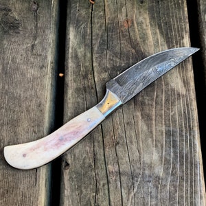 May include: A Damascus steel knife with a curved handle. The blade has a unique pattern, and the handle is off-white with pinkish-brown accents. The knife is set against a weathered wooden surface.