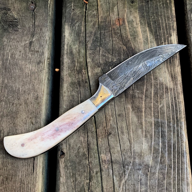 May include: A Damascus steel knife with a curved handle. The blade has a unique pattern, and the handle is off-white with pinkish-brown accents. The knife is set against a weathered wooden surface.
