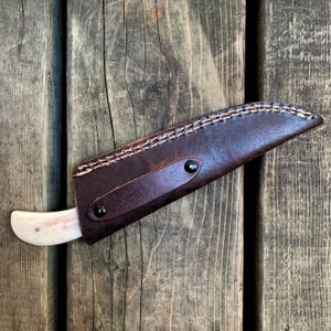May include: A handcrafted knife with a bone handle and a dark brown leather sheath. The sheath is stitched with white thread and secured with two black rivets. The knife is designed for outdoor use.