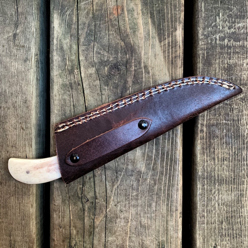 May include: A handcrafted knife with a bone handle and a dark brown leather sheath. The sheath is stitched with white thread and secured with two black rivets. The knife is designed for outdoor use.