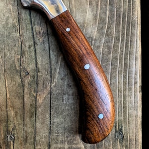 May include: Close-up of a handcrafted knife with a dark brown wooden handle. The handle features three silver rivets and a curved ergonomic design. The blade is partially visible, with a silver and gold finish.