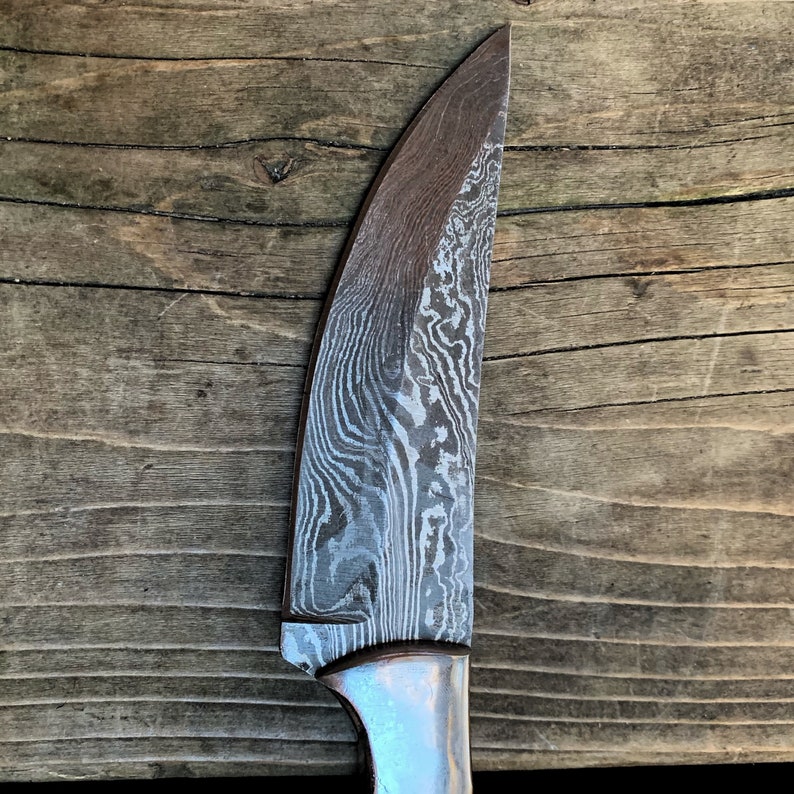 May include: Close-up of a Damascus steel knife blade with a curved edge. The blade has a unique, swirling pattern in shades of gray and silver. The handle is a light silver color. The knife is a tool for cutting.
