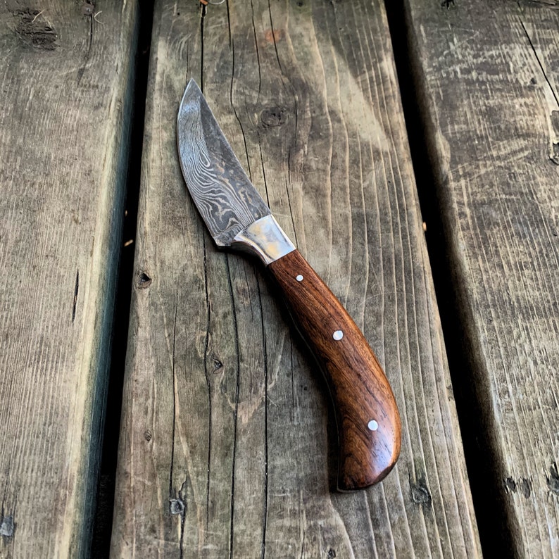 May include: A Damascus steel knife with a curved, dark wood handle. The blade has a unique pattern, and the handle is secured with three silver rivets. The knife is designed for cutting and is suitable for various tasks.