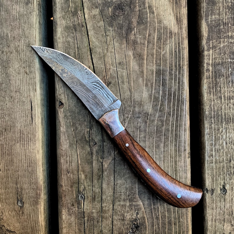 May include: A handcrafted knife with a curved blade and a dark wooden handle. The blade has a unique pattern, and the handle is adorned with small, light-colored accents. The knife rests on a weathered wooden surface.