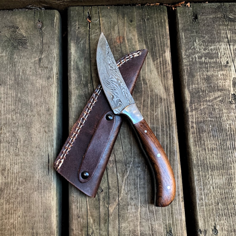 May include: A Damascus steel knife with a wooden handle and a brown leather sheath. The knife blade has a unique pattern, and the handle is dark brown with white accents. The sheath is stitched with white thread and secured with black rivets.