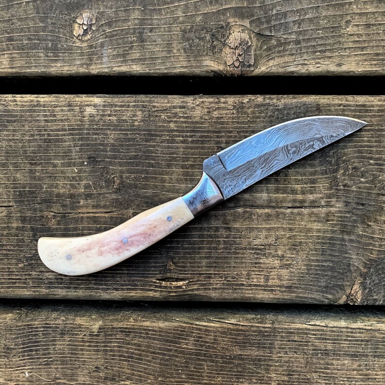 May include: A handcrafted knife with a curved blade and a bone handle. The blade has a unique patterned design. The handle is cream-colored with reddish-brown accents and small blue-grey rivets. The knife is shown on a wooden surface.