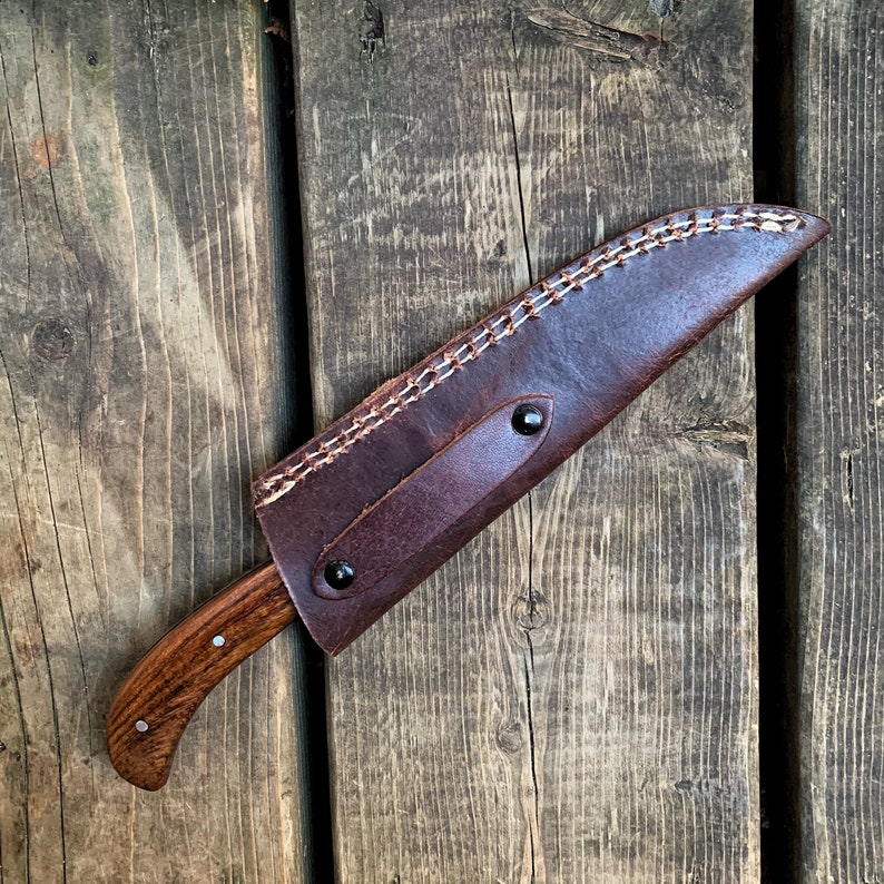 May include: A handcrafted knife with a wooden handle and a leather sheath. The knife blade is partially visible, and the sheath is dark brown with white stitching along the edge. The handle has a curved design and is secured with rivets.