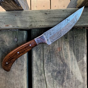May include: A Damascus steel knife with a curved, wooden handle. The blade has a distinctive pattern, and the handle is a dark brown color with silver rivets. The knife is designed for cutting and slicing.