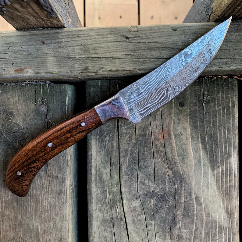 May include: A Damascus steel knife with a curved, wooden handle. The blade has a distinctive pattern, and the handle is a dark brown color with silver rivets. The knife is designed for cutting and slicing.