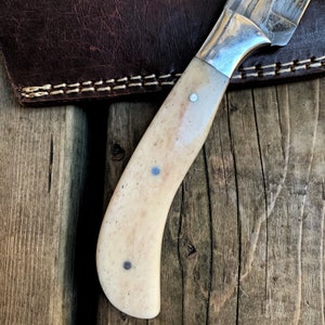 May include: Close-up of a handcrafted knife with a bone handle and a silver bolster. The handle is cream-colored with three small, silver rivets. The blade is partially visible, showing a patterned design. A brown leather sheath is in the background.