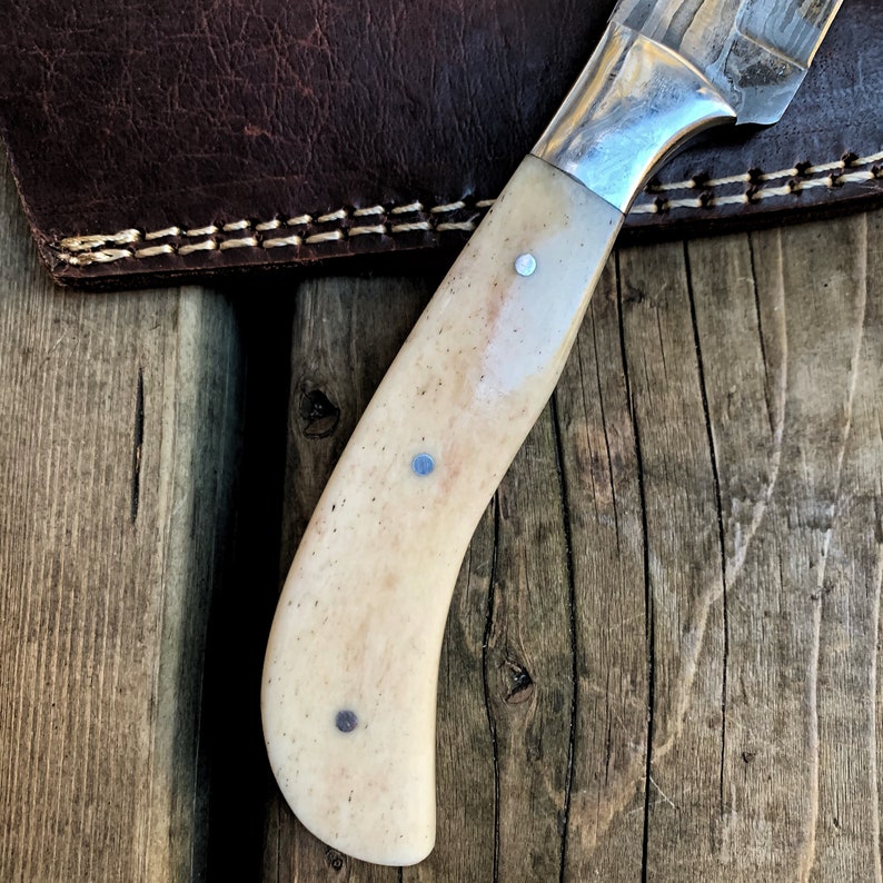 May include: Close-up of a handcrafted knife with a bone handle and a silver bolster. The handle is cream-colored with three small, silver rivets. The blade is partially visible, showing a patterned design. A brown leather sheath is in the background.