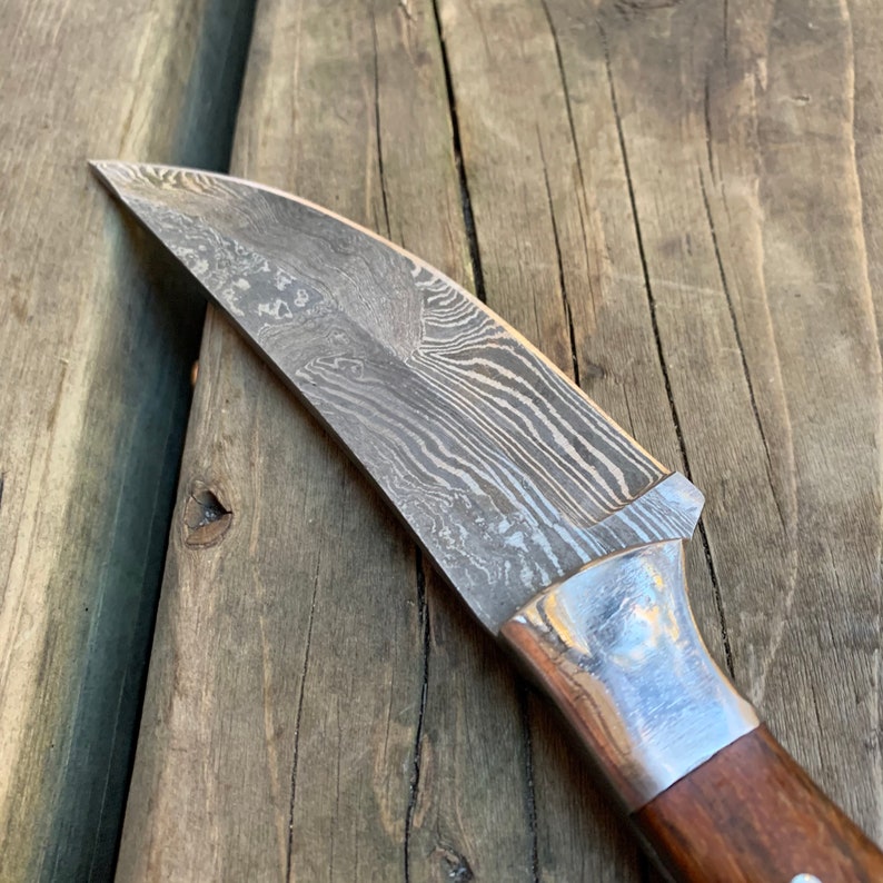 May include: Close-up of a Damascus steel knife blade with a wooden handle. The blade has a unique pattern of swirling lines. The handle is a dark brown color with a silver bolster. The knife is resting on a wooden surface.