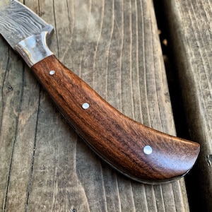May include: Close-up of a Damascus steel knife with a dark brown wooden handle. The handle is adorned with three silver rivets. The blade has a silver bolster and a patterned design.